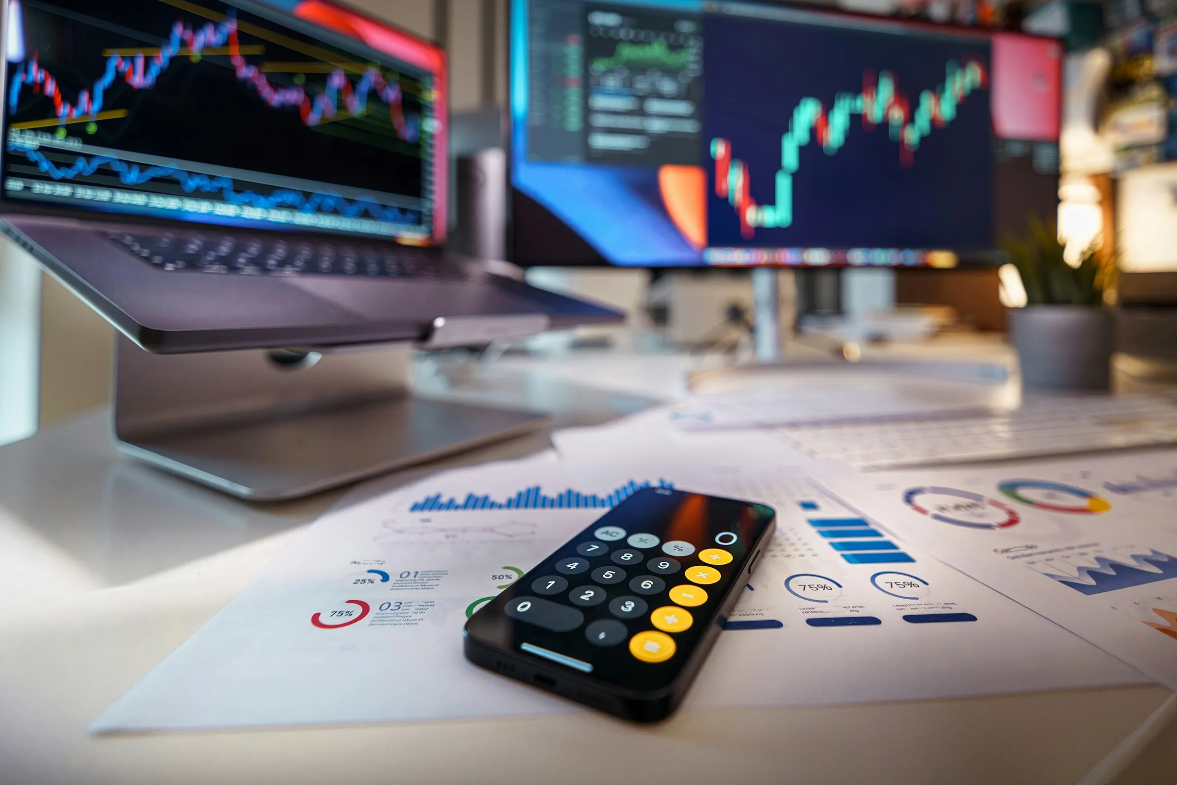A smartphone showing a calculator, lying on printed financial charts in front of a laptop and monitor