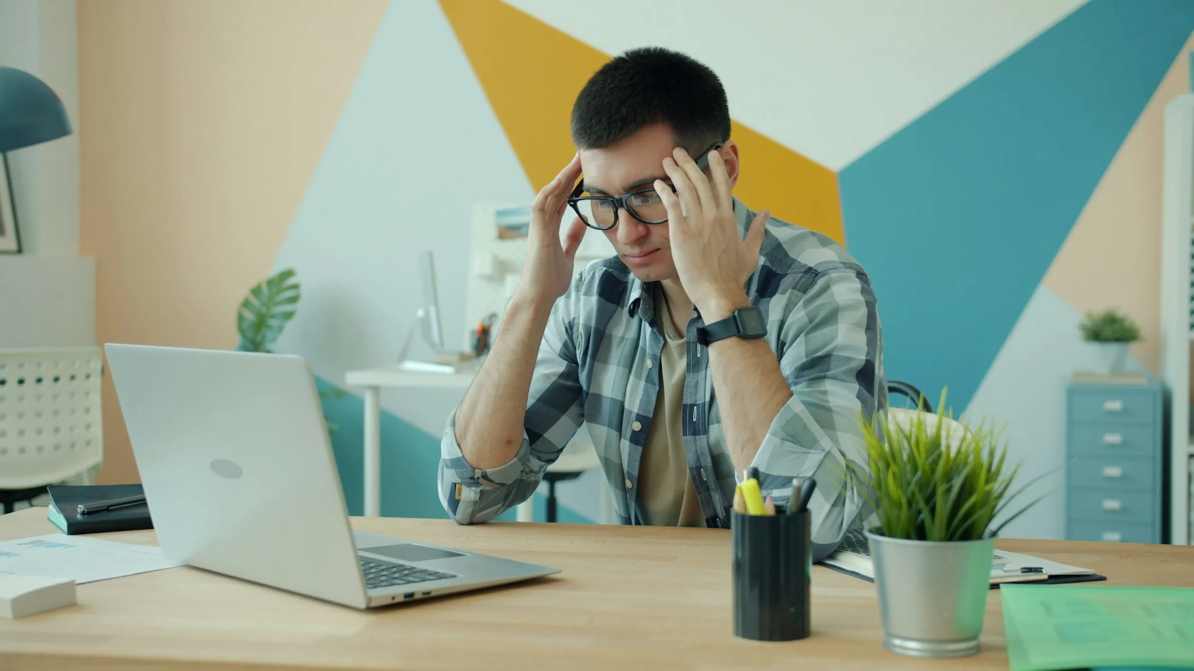 A man at a desk with his hands on his head, looking frustrated at his laptop